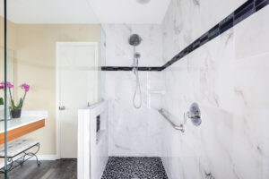A modern bathroom with a large walk-in shower featuring white marble walls and a pebble-tiled floor. The shower includes a handheld showerhead and a safety grab bar. Nearby is a wooden vanity with pink orchids in a glass vase.