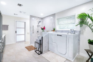 Spacious, modern bathroom with a walk-in tub, gray cabinets, and a marble-tiled shower. A potted plant is in the corner, and a window with blinds offers natural light. Towels hang on racks, and decor includes flowers and a small rug.