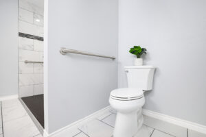 A modern bathroom with light gray walls features a toilet next to a potted plant on the tank and a metal grab bar on the wall. A tiled walk-in shower with a glass wall is adjacent. The floor is white tile with subtle gray veining.