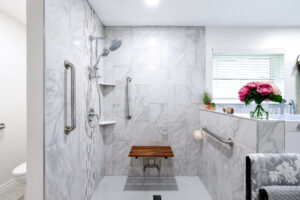 A modern bathroom with a marble shower, featuring a wooden fold-down bench and stainless steel grab bars. A small window with blinds and a ledge with pink flowers, candles, and a plant add a decorative touch.