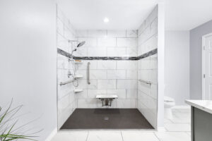 Spacious shower with a white marble tile design, featuring grab bars, a handheld showerhead, and a foldable seat. Dark accent tiles form a horizontal stripe. A toilet is visible nearby, and a green plant adds a touch of color.