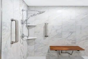 A modern shower with marbled tiles, a handheld showerhead on an adjustable rail, two white shelves, stainless steel grab bars, and a foldable wooden bench.
